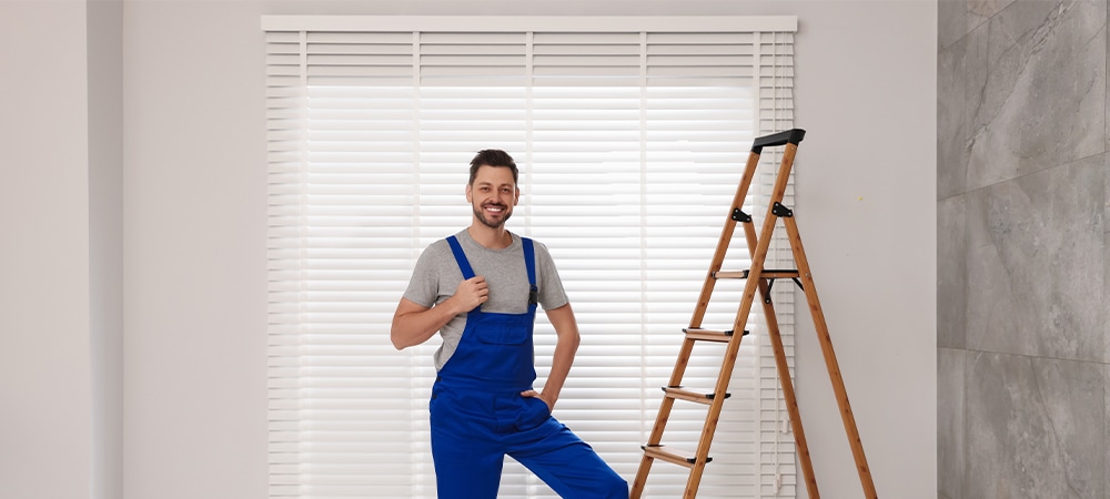 Worker in uniform and stepladder near horizontal window blinds indoors