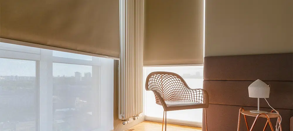 Modern automatic roller blinds and sun shades cover large windows, diffusing natural light into cozy room. Blackout and screen fabrics close-up. Space features stylish chair and minimalist side table.
