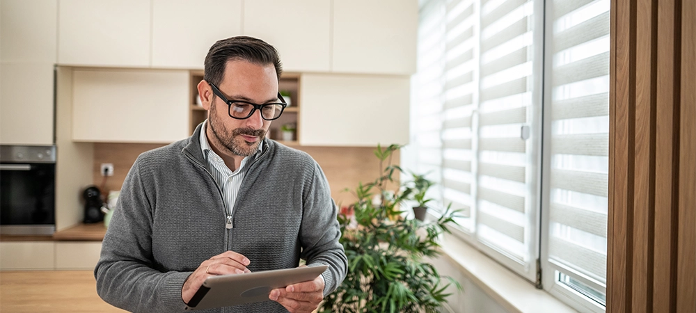 Man using a digital tablet to manage smart home automation features like window blinds from a modern kitchen, representing convenience and technological integration