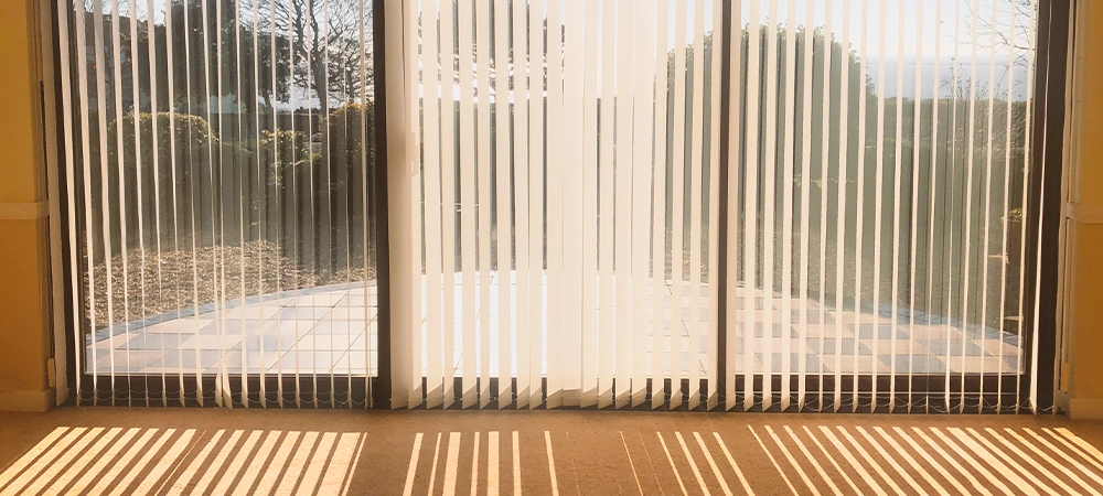 sunlight shining through full length white vertical blinds in front of three glass sliding french doors leading to a patio, garden and sea view, creating shadow stripes.