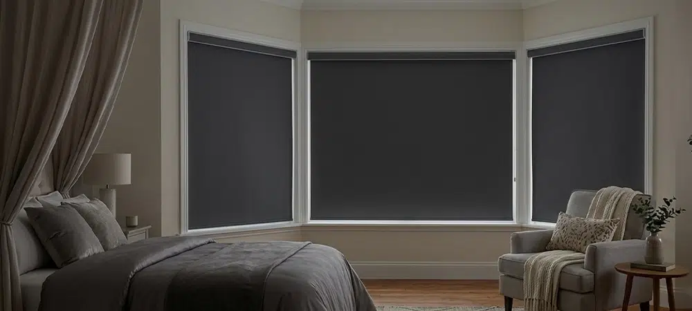 interior view of a cozy bedroom showing a large bay window covered with dark grey blackout roller shades to completely block out external light.