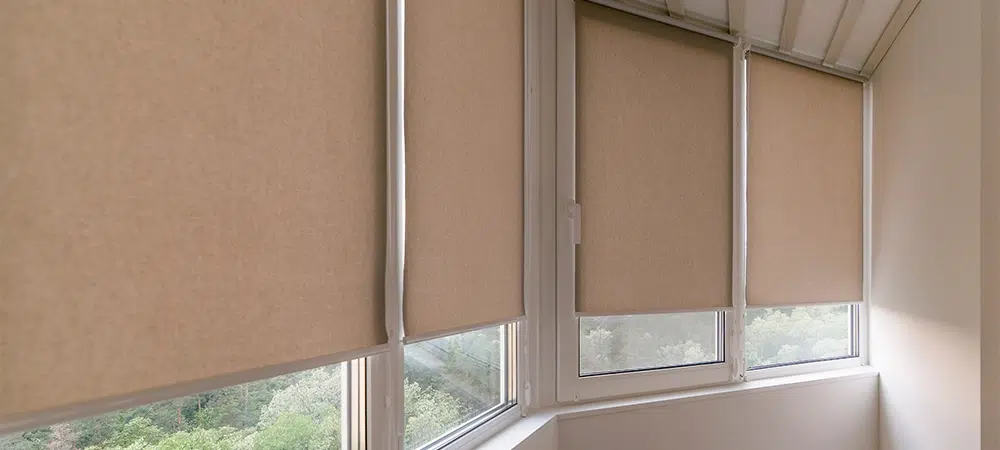 Modern roller blinds in a neutral beige color covering windows in a bright enclosed balcony space. Natural light filters through roller shades, with a greenery outside, creating a cozy atmosphere.